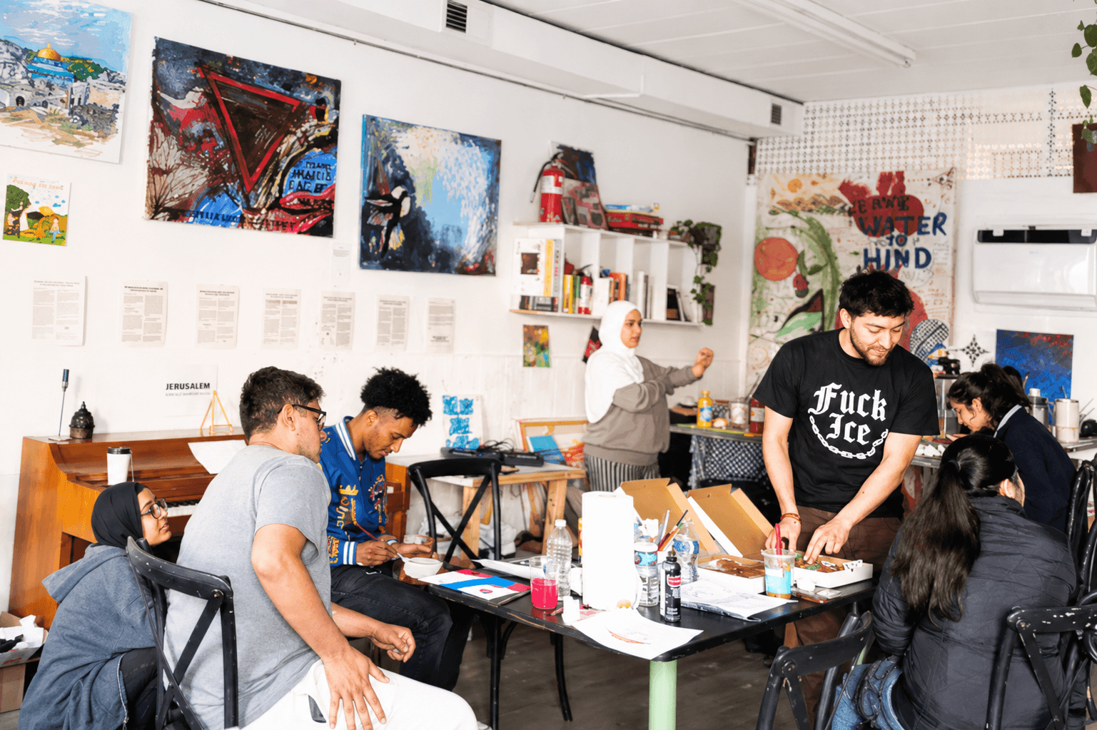 Young people painting and making art together around tables in the academy's storefront studio, its walls covered with student paintings and a Love Letter to Hind mural